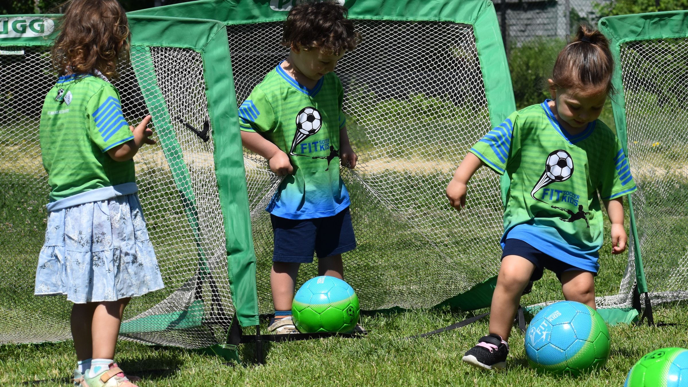 Three young children in green Fit Soccer Kids jerseys playing with soccer balls beside pop-up goals on a sunny field