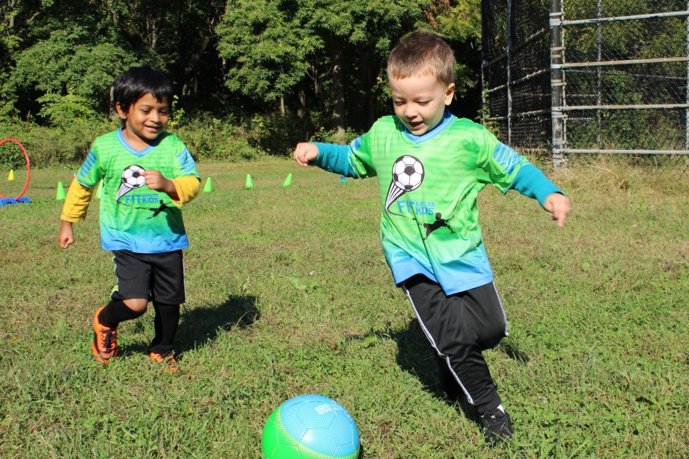 Two young Fit Soccer Kids players in green jerseys running after a soccer ball on a grass field