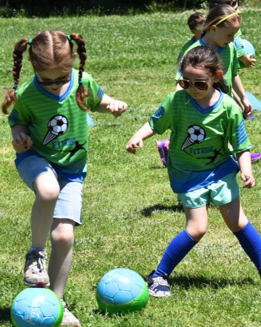 Two young Fit Soccer Kids players dribbling soccer balls together on the field